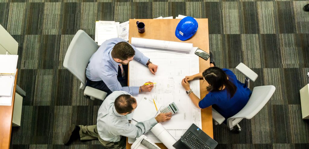 A top down view of three people in business attire sitting around a table looking at a large office plan schematic